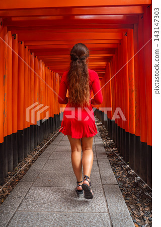 A lone visitor in a red dress walks through Senbon Torii at Fushimi Inari Taisha in Kyoto, Japan. Repeating gates form a vertical, centered corridor of color and shadow. A lone visitor in a red dress walks through Senbon Torii at Fushimi Inari Taisha in Kyoto, Japan. Repeating gates form a vertical, centered corridor of color and shadow. 143147303
