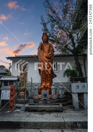 A bronze toned Kannon stands with smaller figures and prayer beads in a Kyoto temple courtyard. Warm sunset light highlights flowing robes and a calm mudra. 143147366