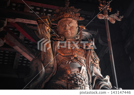 A carved temple guardian stands in warm light, gripping a staff and raising a small pagoda in a dim hall in Nara, Japan, with heavy beams and latticework overhead. A carved temple guardian stands in warm light, gripping a staff and raising a small pagoda in a dim hall in Nara, Japan, with heavy beams and latticework overhead. 143147446