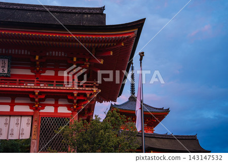 A vermilion temple gate and tiered pagoda stand at Kiyomizu dera in Kyoto at dusk. Warm lights reveal eaves, latticework, and a Japanese flag in soft blue light. A vermilion temple gate and tiered pagoda stand at Kiyomizu dera in Kyoto at dusk. Warm lights reveal eaves, latticework, and a Japanese flag in soft blue light. 143147532