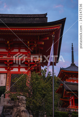 Vermilion temple buildings and pagoda rise at Kiyomizu dera in Kyoto, Japan. A Japanese flag and stone guardian statues stand in the foreground in early evening light. 143147536