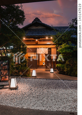 A Kyoto machiya teahouse glows at dusk, with noren, tiled roof, and wood lattice. Lanterns line a stone path over raked white gravel, leading to a warm doorway. A Kyoto machiya teahouse glows at dusk, with noren, tiled roof, and wood lattice. Lanterns line a stone path over raked white gravel, leading to a warm doorway. 143147619
