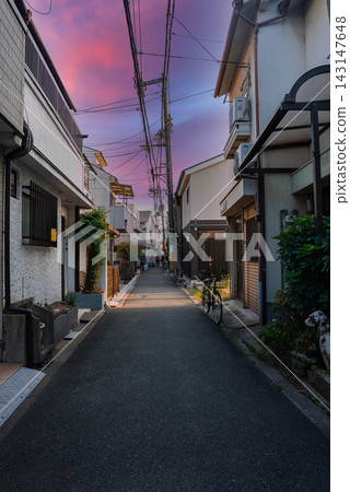 A narrow Osaka alley at dusk shows compact houses, porch lights, signs, and a bicycle by a doorway, with power lines converging at a tall utility pole. A narrow Osaka alley at dusk shows compact houses, porch lights, signs, and a bicycle by a doorway, with power lines converging at a tall utility pole. 143147648