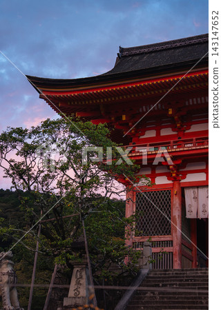 Kiyomizu dera Nio mon in Kyoto, Japan stands at dusk, wooden brackets and lattice windows glow, stone steps lead past guardian statues, a leafy tree frames the view. 143147652