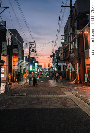 Low rise shops and restaurants in Nara at dusk display neon signs and lanterns. Overhead lines cross as pedestrians and a dog walker move along wet paving. Low rise shops and restaurants in Nara at dusk display neon signs and lanterns. Overhead lines cross as pedestrians and a dog walker move along wet paving. 143147682