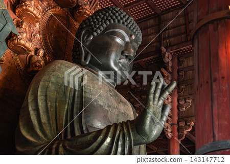 A towering bronze Buddha fills a vast wooden hall in Nara, Japan, right hand raised in blessing, golden mandorla and timber beams visible, warm light emphasizes scale and craft. A towering bronze Buddha fills a vast wooden hall in Nara, Japan, right hand raised in blessing, golden mandorla and timber beams visible, warm light emphasizes scale and craft. 143147712