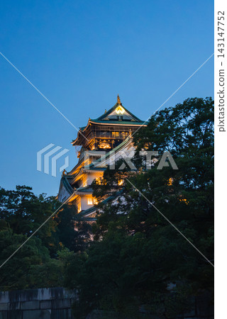 Osaka Castle in Osaka, Japan rises above dark trees at dusk. Green tiles, gold accents, and ornate gables stand out as warm lights glow against deep blue. Osaka Castle in Osaka, Japan rises above dark trees at dusk. Green tiles, gold accents, and ornate gables stand out as warm lights glow against deep blue. 143147752
