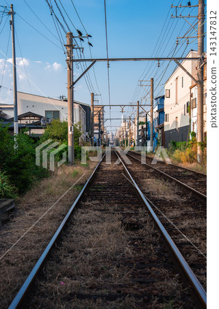 Ground level view shows narrow gauge tracks through an Osaka neighborhood, with utility poles, catenary wires, and houses. Late afternoon light casts long shadows. Ground level view shows narrow gauge tracks through an Osaka neighborhood, with utility poles, catenary wires, and houses. Late afternoon light casts long shadows. 143147812