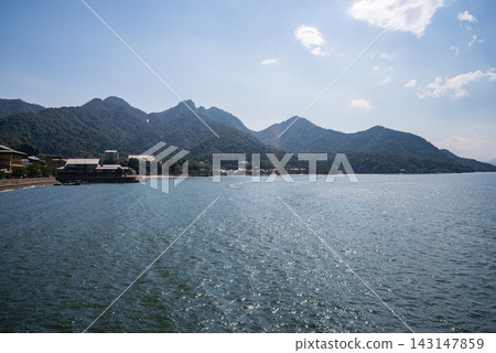 Wide view of the Seto Inland Sea frames Mount Misen on Itsukushima Island near Hiroshima. Gentle waves lead to a coastal settlement with traditional buildings along the shore. Wide view of the Seto Inland Sea frames Mount Misen on Itsukushima Island near Hiroshima. Gentle waves lead to a coastal settlement with traditional buildings along the shore. 143147859