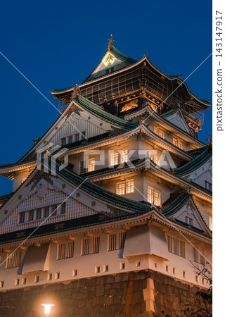 Osaka Castle in Osaka, Japan stands at dusk, green tiled roofs and gilded details lit warm, stone walls below, ornate gables and white plaster seen in soft light 143147917