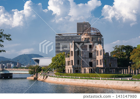 The Hiroshima Peace Memorial stands by the Motoyasu River in Hiroshima, Japan. People stroll along the promenade as modern buildings and distant hills frame the ruin. The Hiroshima Peace Memorial stands by the Motoyasu River in Hiroshima, Japan. People stroll along the promenade as modern buildings and distant hills frame the ruin. 143147980