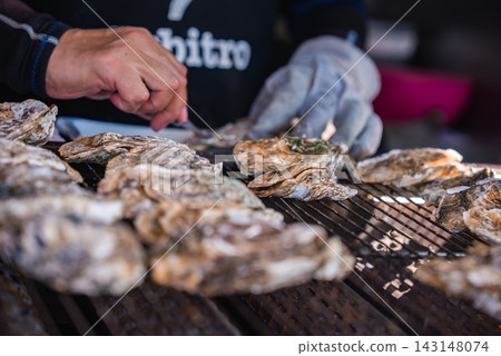 Two people grill and shuck large oysters on a metal grate on Itsukushima near Hiroshima, Japan. Close hands, one gloved, work rows of rough shells in tight focus. Two people grill and shuck large oysters on a metal grate on Itsukushima near Hiroshima, Japan. Close hands, one gloved, work rows of rough shells in tight focus. 143148074