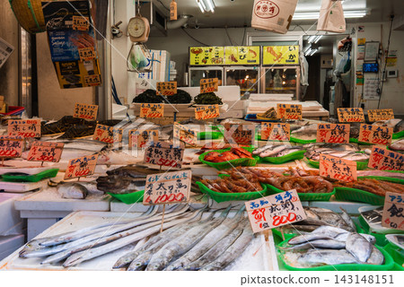 A Tokyo market stall displays fish, prawns, squid, and shellfish on green trays with handwritten Japanese price tags, scales and hanging signs visible in the background. A Tokyo market stall displays fish, prawns, squid, and shellfish on green trays with handwritten Japanese price tags, scales and hanging signs visible in the background. 143148151