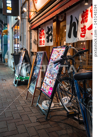 A narrow Tokyo street shows small restaurants with white noren, glowing menu boards, parked bicycles, handwritten signs, tiled pavement, and casual evening walkers. A narrow Tokyo street shows small restaurants with white noren, glowing menu boards, parked bicycles, handwritten signs, tiled pavement, and casual evening walkers. 143148194