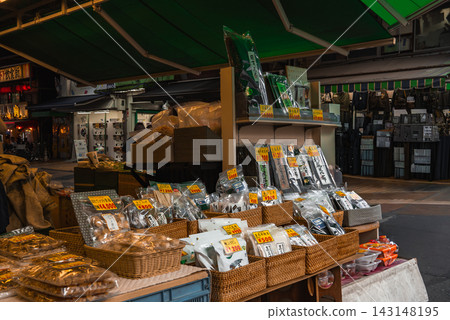 A Tokyo shotengai stall shows baskets and shelves of packaged snacks and dried goods with bright yellow yen tags, neighboring shops and awnings visible as shoppers browse. A Tokyo shotengai stall shows baskets and shelves of packaged snacks and dried goods with bright yellow yen tags, neighboring shops and awnings visible as shoppers browse. 143148195
