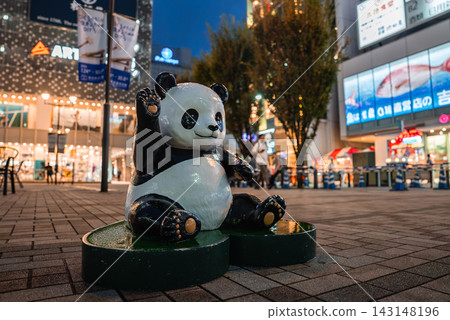 A black and white panda statue sits and waves in a Tokyo shopping district, with lit storefronts, billboards with Japanese text, and tree lined walkways in early evening. A black and white panda statue sits and waves in a Tokyo shopping district, with lit storefronts, billboards with Japanese text, and tree lined walkways in early evening. 143148196