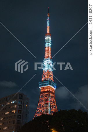 Tokyo Tower stands in Minato, Tokyo at night, its orange and blue lights glowing. Nearby apartments and treetops frame the tower from a close street level view. Tokyo Tower stands in Minato, Tokyo at night, its orange and blue lights glowing. Nearby apartments and treetops frame the tower from a close street level view. 143148207