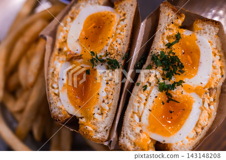 A close up of a Japanese tamago sando in Tokyo shows creamy egg salad, halved soft boiled eggs, chopped herbs, and pepper in a paper sleeve beside golden fries. A close up of a Japanese tamago sando in Tokyo shows creamy egg salad, halved soft boiled eggs, chopped herbs, and pepper in a paper sleeve beside golden fries. 143148208