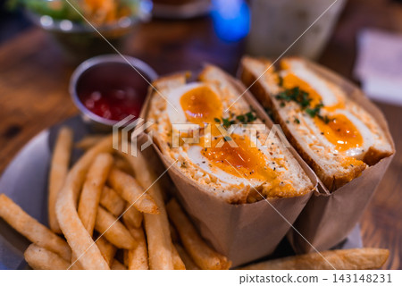 Close view of a halved Japanese egg sandwich with jammy yolks, herbs, thick fries, and ketchup in Tokyo, warm tones and shallow depth of field emphasize texture. Close view of a halved Japanese egg sandwich with jammy yolks, herbs, thick fries, and ketchup in Tokyo, warm tones and shallow depth of field emphasize texture. 143148231