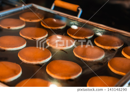Golden brown dorayaki pancakes puff as they cook on a large griddle at a Tokyo street market, even rows on a shiny metal surface suggest a vendor setup. 143148290