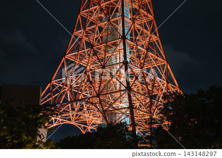 Illuminated lattice base of Tokyo Tower rises behind dark trees in Minato, Tokyo. Orange steel beams and deck supports stand out in a close night view. Illuminated lattice base of Tokyo Tower rises behind dark trees in Minato, Tokyo. Orange steel beams and deck supports stand out in a close night view. 143148297