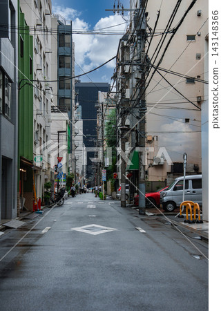 A Tokyo side street runs between apartments and shops, with power lines, wet pavement, scooters, vans, green awnings, and vending machines in a central district scene. A Tokyo side street runs between apartments and shops, with power lines, wet pavement, scooters, vans, green awnings, and vending machines in a central district scene. 143148366