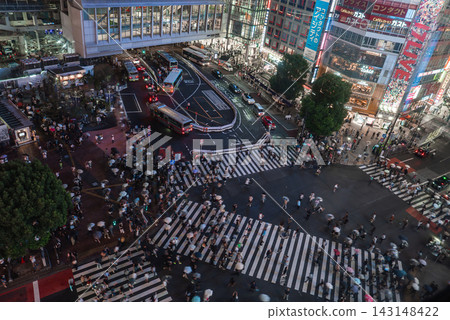 Hundreds cross the diagonal crosswalks at Shibuya Crossing in Tokyo, umbrellas moving past neon storefronts and billboards as traffic lines buses by Shibuya Station. 143148422