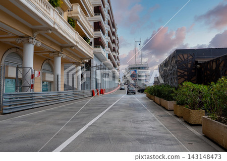 Soft light reflects on Monaco harbor balconies and a lattice pavilion as cars drive past crash barriers and red bollards toward Port Hercules with yachts and masts. 143148473