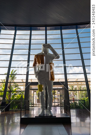A white astronaut statue with gold fabric and bag stands in a glass atrium in Nice, France. Tall slatted windows reveal sky and palm trees, shot low with soft daylight. A white astronaut statue with gold fabric and bag stands in a glass atrium in Nice, France. Tall slatted windows reveal sky and palm trees, shot low with soft daylight. 143148483