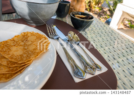 Thin lacy crepes rest on a white plate beside silverware on a napkin, with small bowls of jam and a pepper mill on a wicker table in sunlit Antibes, France. Thin lacy crepes rest on a white plate beside silverware on a napkin, with small bowls of jam and a pepper mill on a wicker table in sunlit Antibes, France. 143148487
