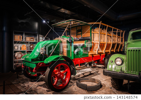 An indoor auto museum display presents a restored early 1900s truck with green hood, red chassis, wooden cargo body, and brass fittings under low key lighting An indoor auto museum display presents a restored early 1900s truck with green hood, red chassis, wooden cargo body, and brass fittings under low key lighting 143148574