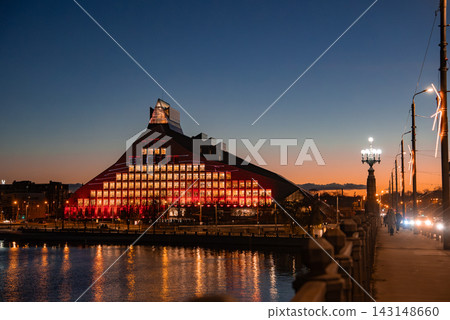 Riga, Latvia, dusk view of the National Library of Latvia glowing red, windows form a grid, reflections on the Daugava River, streetlights and traffic move along the embankment Riga, Latvia, dusk view of the National Library of Latvia glowing red, windows form a grid, reflections on the Daugava River, streetlights and traffic move along the embankment 143148660