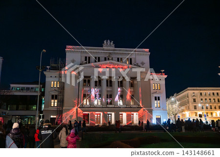 Latvian National Opera in Riga glows at night as red and white light maps the facade. Crowds in winter coats watch Staro Riga 2025 amid historic architecture. Latvian National Opera in Riga glows at night as red and white light maps the facade. Crowds in winter coats watch Staro Riga 2025 amid historic architecture. 143148661