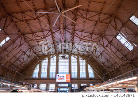 Daytime view inside Riga Central Market in Latvia shows arched windows, steel trusses, and a large ceiling fan above vendor stalls with light on a digital sign. Daytime view inside Riga Central Market in Latvia shows arched windows, steel trusses, and a large ceiling fan above vendor stalls with light on a digital sign. 143148671