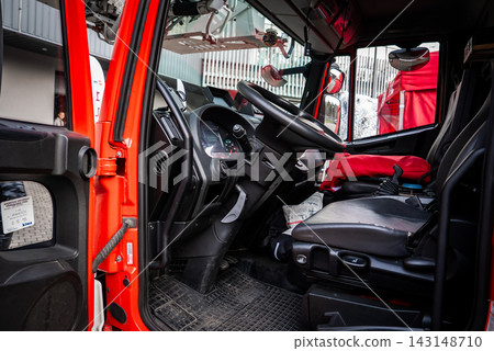 Open driver cabin of a red fire truck shows wheel, gauges, rugged mats, black captain seat, radio mic, mirrors, and gear in a museum display under natural indoor light. Open driver cabin of a red fire truck shows wheel, gauges, rugged mats, black captain seat, radio mic, mirrors, and gear in a museum display under natural indoor light. 143148710