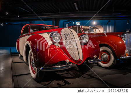 A red and cream vintage coupe stands under museum lighting, with sweeping fenders, teardrop headlights, and a tall chromed grille, classic cars lined on polished floors. A red and cream vintage coupe stands under museum lighting, with sweeping fenders, teardrop headlights, and a tall chromed grille, classic cars lined on polished floors. 143148738
