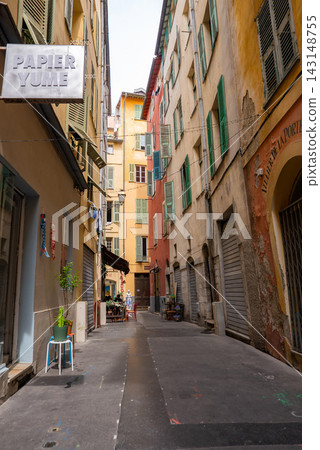 A narrow alley in Vieux Nice, France, leads to a tiny cafe terrace. Ochre and pastel walls, green shutters, arched doors, metal roll downs, and PAPIER YUME appear. A narrow alley in Vieux Nice, France, leads to a tiny cafe terrace. Ochre and pastel walls, green shutters, arched doors, metal roll downs, and PAPIER YUME appear. 143148755