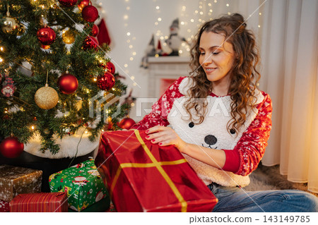 Smiling woman opening a red Christmas gift by decorated tree Smiling woman opening a red Christmas gift by decorated tree 143149785