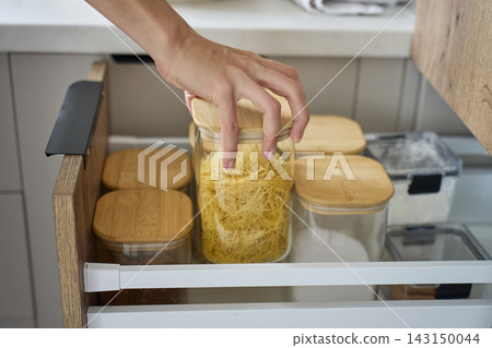 Hand taking glass jar filled with dry pasta from open kitchen drawer Hand taking glass jar filled with dry pasta from open kitchen drawer 143150044