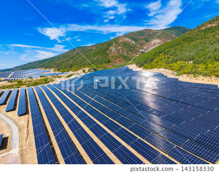 Solar panels cover a large area in a sunny landscape near mountains with a clear blue sky above Solar panels cover a large area in a sunny landscape near mountains with a clear blue sky above 143158330