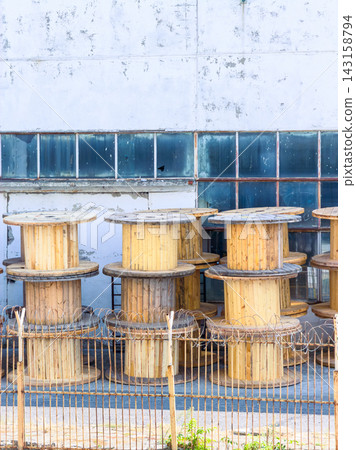 Large wooden spools lined up next to a building in an industrial area during daylight Large wooden spools lined up next to a building in an industrial area during daylight 143158794