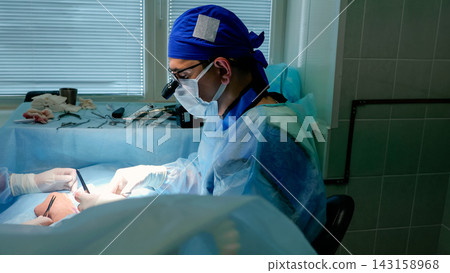 Surgeon performing delicate procedure in sterile operating room with masked medical team, bright surgical lighting, blue drapes, protective eyewear, gloves, and prepared instruments nearby Surgeon performing delicate procedure in sterile operating room with masked medical team, bright surgical lighting, blue drapes, protective eyewear, gloves, and prepared instruments nearby 143158968
