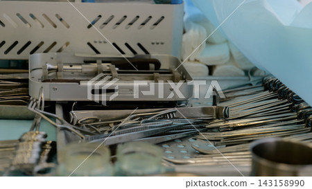 Sterile dental instruments arranged on clinical tray with metal cassette, forceps, mirrors, probes, glass cups, and blue protective equipment in clean treatment room setting Sterile dental instruments arranged on clinical tray with metal cassette, forceps, mirrors, probes, glass cups, and blue protective equipment in clean treatment room setting 143158990