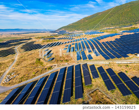 Large solar panel farm on a hillside in a rural area with blue sky and mountains in the background in the daytime 143159025