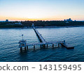 Sunset over a pier with people walking near water and buildings in the background 143159495
