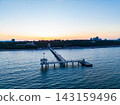 View of a pier extending into the water during sunset with buildings in the background and a clear sky 143159496