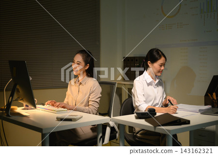 Two Businesswomen Working Late at Night in Office with Data Charts Projected on Wall Two Businesswomen Working Late at Night in Office with Data Charts Projected on Wall 143162321