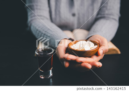 Person holding a wooden bowl of communion wafers beside a dark red beverage and open book, illustrating Christian worship, sacred sharing, prayer, faith, remembrance, and spiritual devotion. Person holding a wooden bowl of communion wafers beside a dark red beverage and open book, illustrating Christian worship, sacred sharing, prayer, faith, remembrance, and spiritual devotion. 143162415