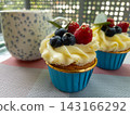 Delicious cupcake with cream frosting, blueberries, and raspberries in a blue wrapper placed on a woven outdoor table next to a ceramic coffee mug. Soft natural light and bokeh background create 143166292