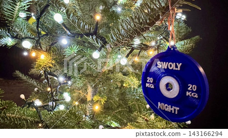 A blue weight plate ornament hangs on a Christmas tree surrounded by warm festive lights. The sporty decoration blends holiday cheer with gym inspiration, creating a unique fitness themed New Year A blue weight plate ornament hangs on a Christmas tree surrounded by warm festive lights. The sporty decoration blends holiday cheer with gym inspiration, creating a unique fitness themed New Year 143166294